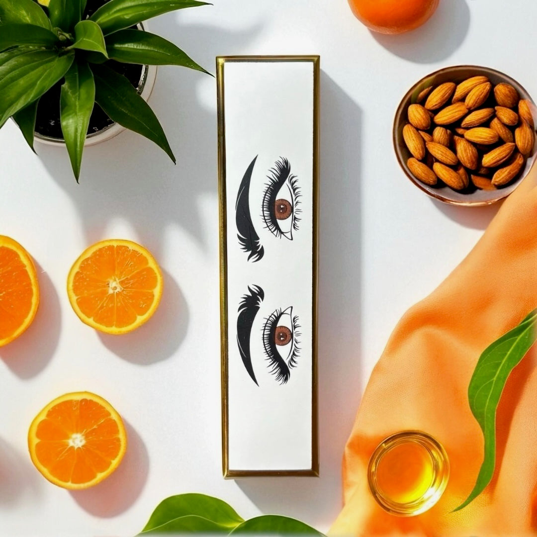 halva beauty nourishing brow oil packaging with eye and brow illustration, styled with fresh orange slices, almonds, plant leaves, orange fabric, and glass bowl of oil.