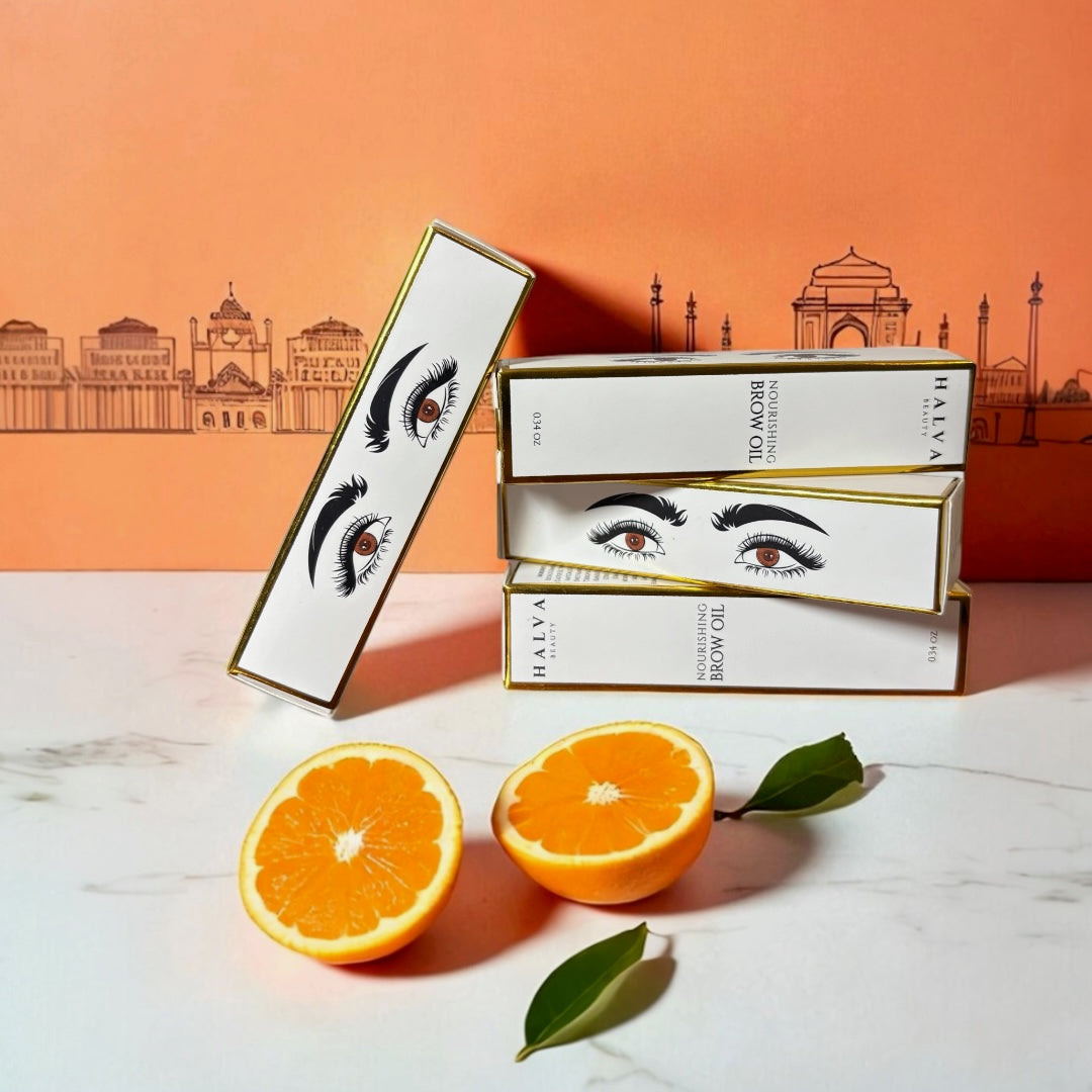 halva beauty nourishing brow oil packaging with eye and brow illustration, stacked against illustrated orange background of indian monuments, styled with fresh orange halves and green leaves on marble surface.
