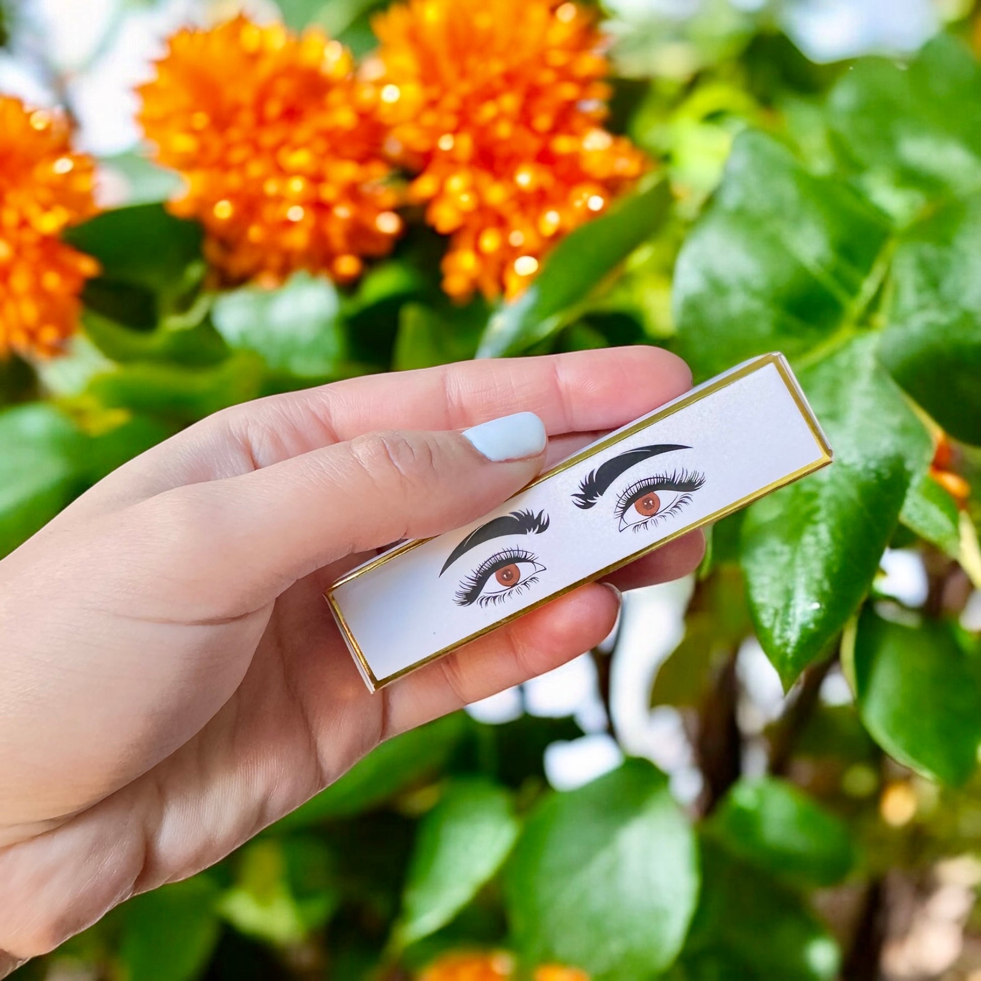 halva beauty nourishing brow oil packaging with illustrated eyes design held in hand against vibrant orange flowers and green leaves background