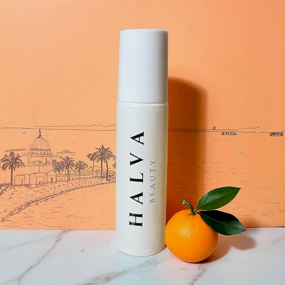 halva beauty nourishing brow oil packaging tube displayed with fresh orange and green leaves, set against illustrated orange background with palm trees and domed building.