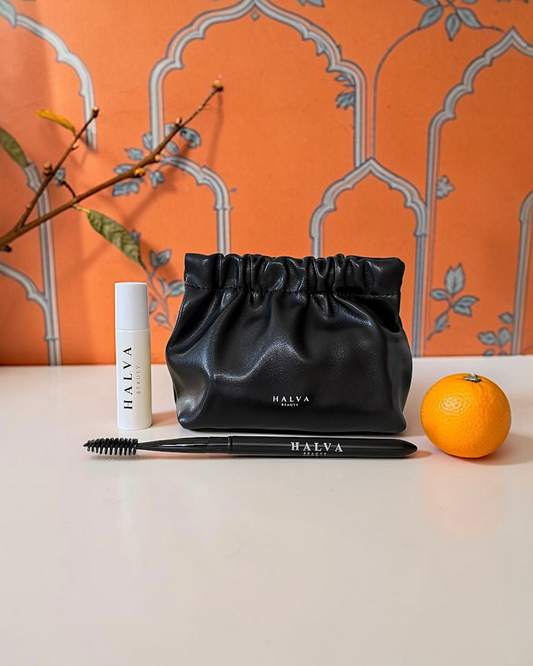 Black Halva Beauty branded cosmetic pouch that shows the Halva Beauty Nourishing Brow Oil and Halva Beauty Spoolie next to it. The products are surrounded by an orange, branches and an Indian-inspired orange wallpaper.