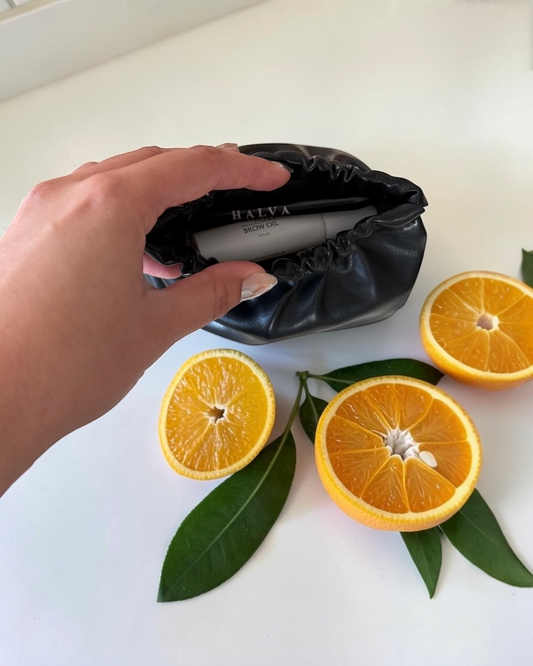 Hand holding a black cosmetic pouch with Halva branding, that shows the Halva Beauty Nourishing Brow Oil and Halva Beauty Spoolie inside, surrounded by sliced oranges and leaves on a white surface.