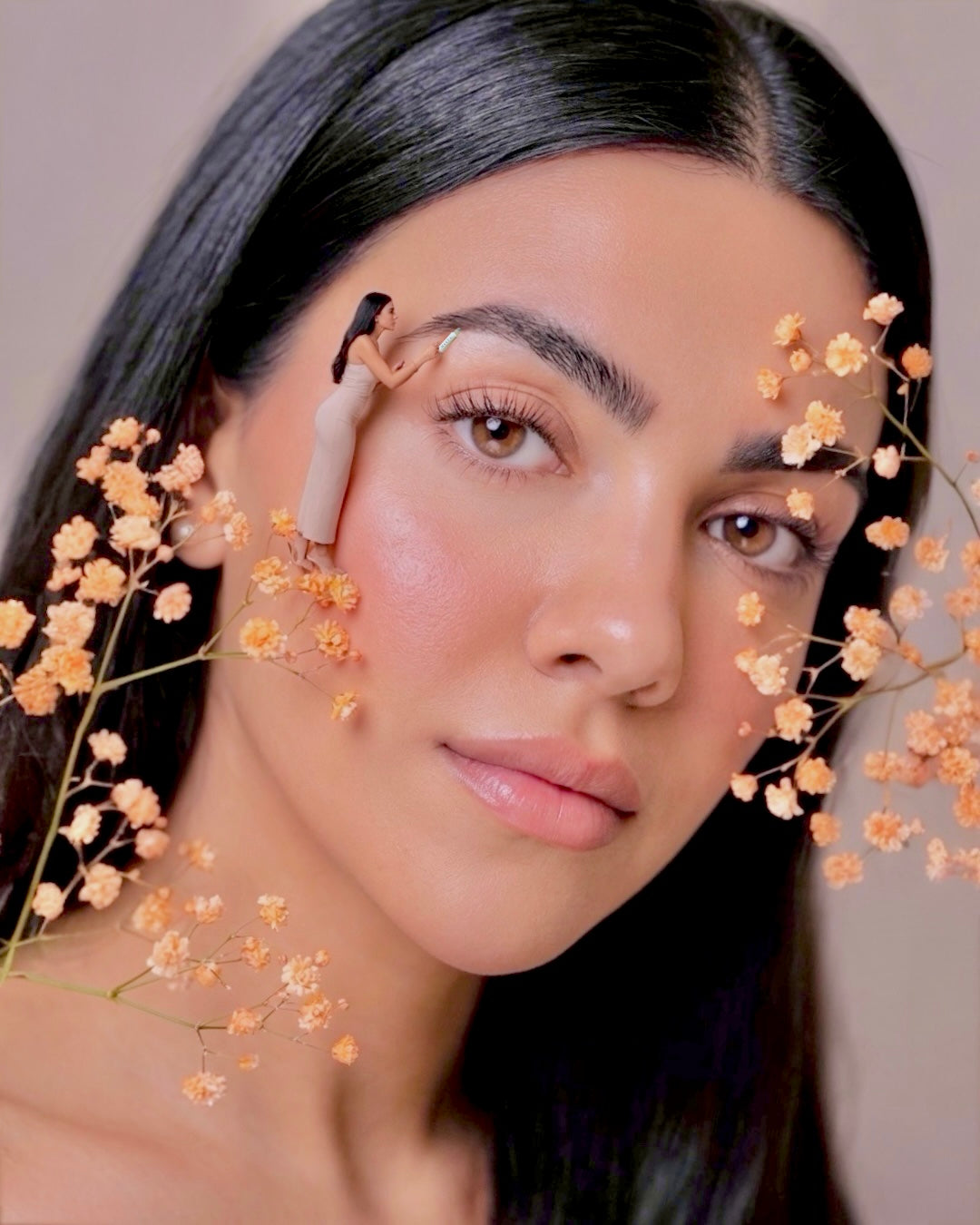 Halva beauty founder with a miniature version of herself applying the halva beauty nourishing brow oil, with flowers around her face against a neutral background