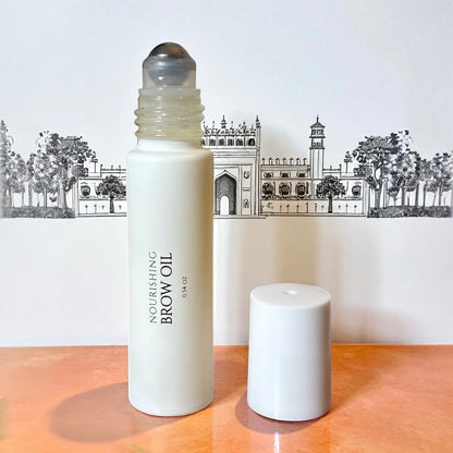 halva beauty nourishing brow oil bottle with rollerball applicator and white cap, displayed against illustrated indian architecture background on peach surface.